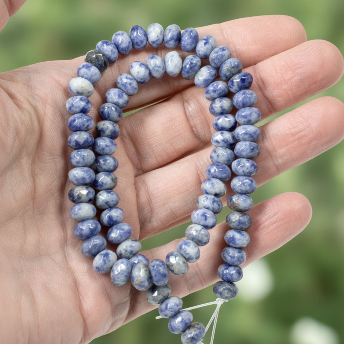 Hackberry Creek faceted denim lapis rondelle beads in blue and gray held in a hand against a blurred green background.