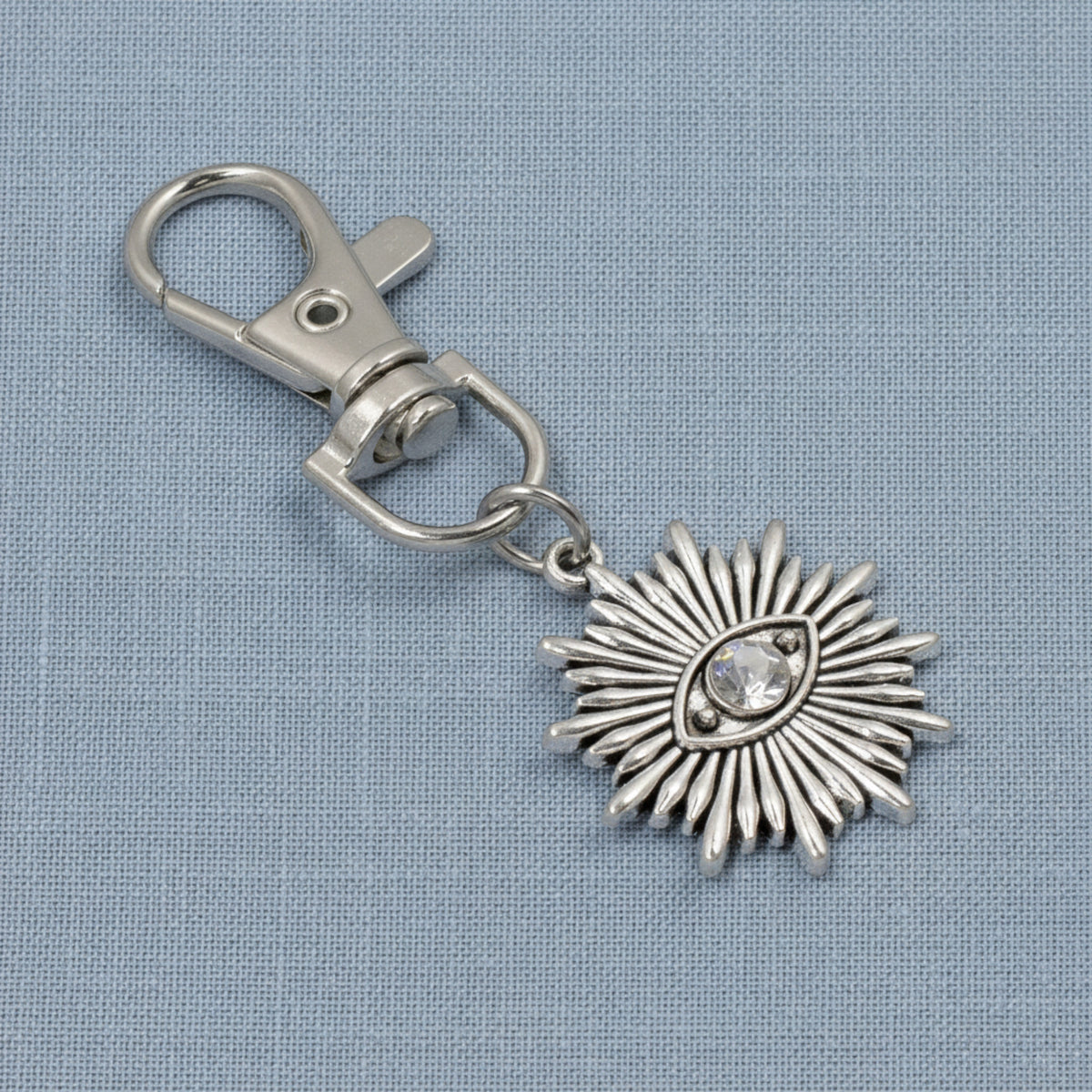 Hackberry Creek silver metal alloy evil eye sunburst bag charm with a clear rhinestone accent on a blue fabric background