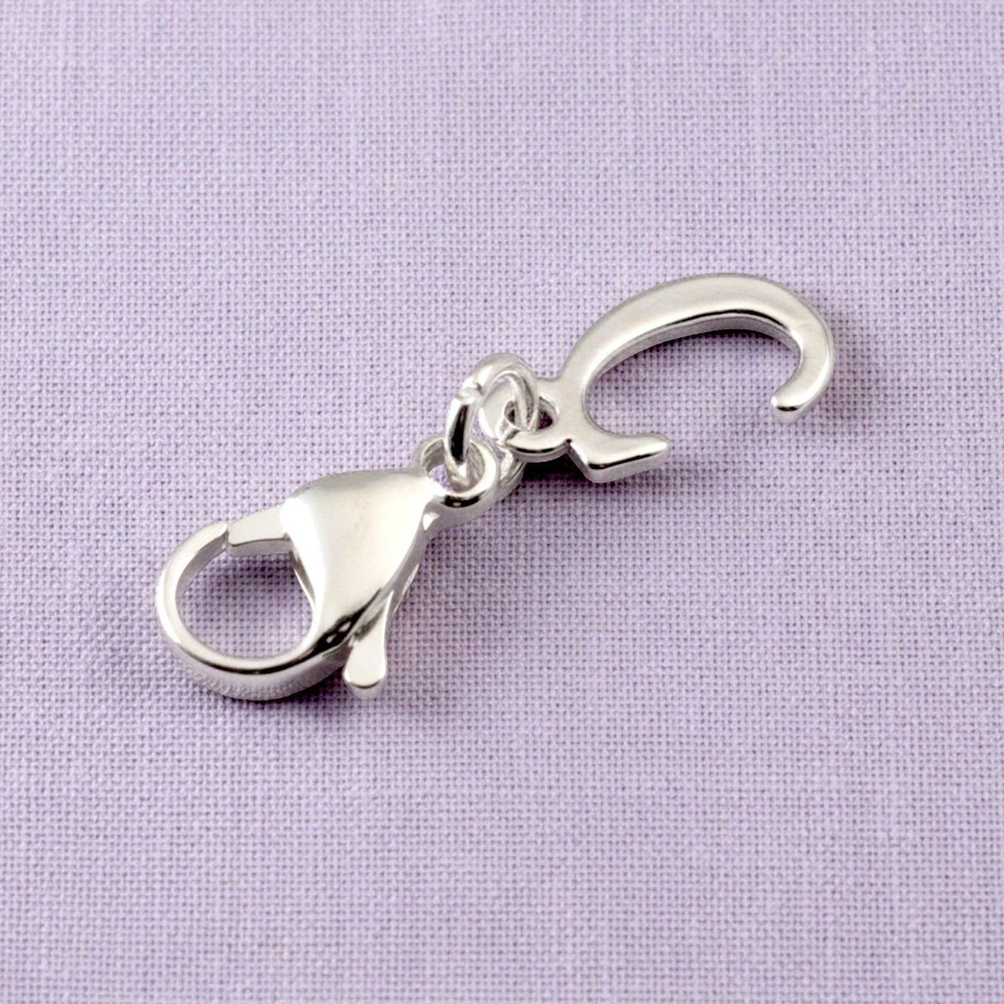 Hackberry Creek silver cursive letter C charm with a stainless steel lobster clasp on a textured lavender background.