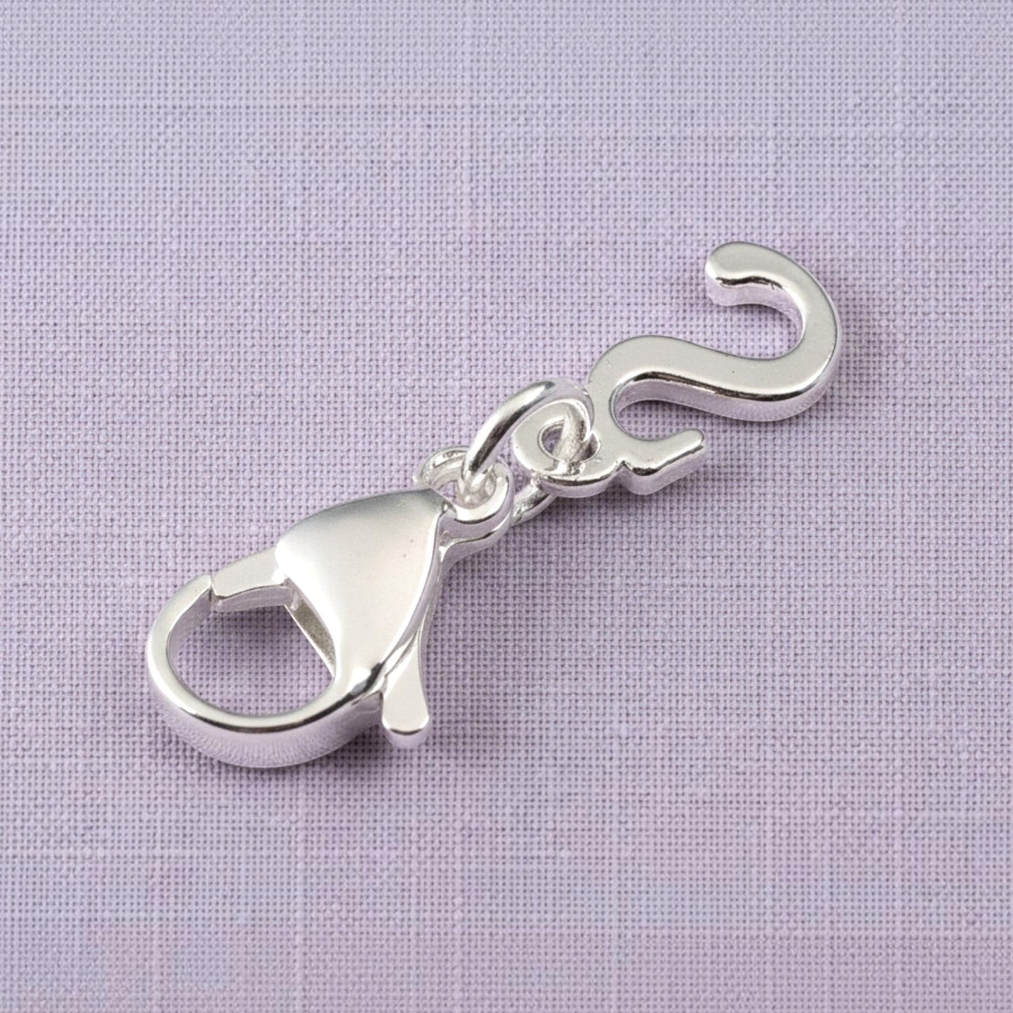 Hackberry Creek silver cursive letter S charm with a stainless steel lobster clasp on a textured purple background.