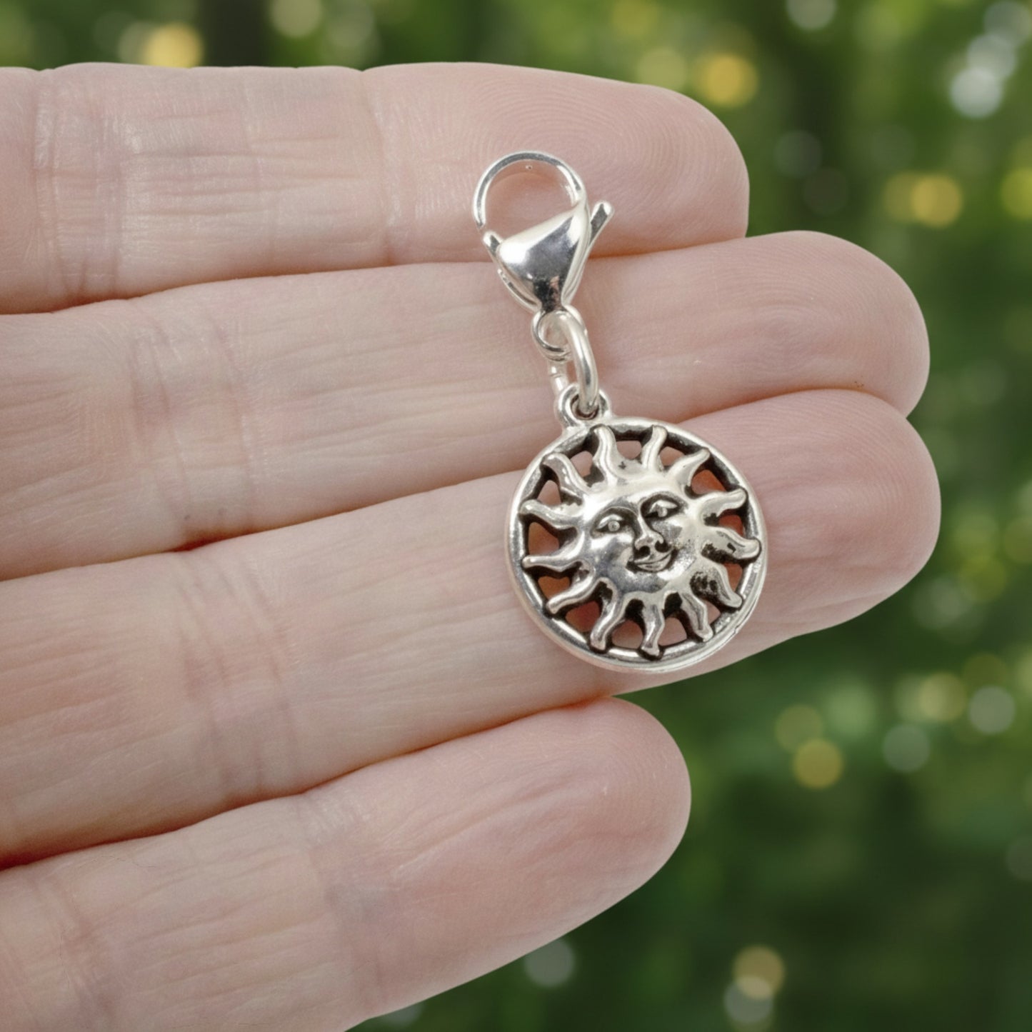 Hackberry Creek silver sun charm with a lobster clasp held in a hand against a blurred green background.
