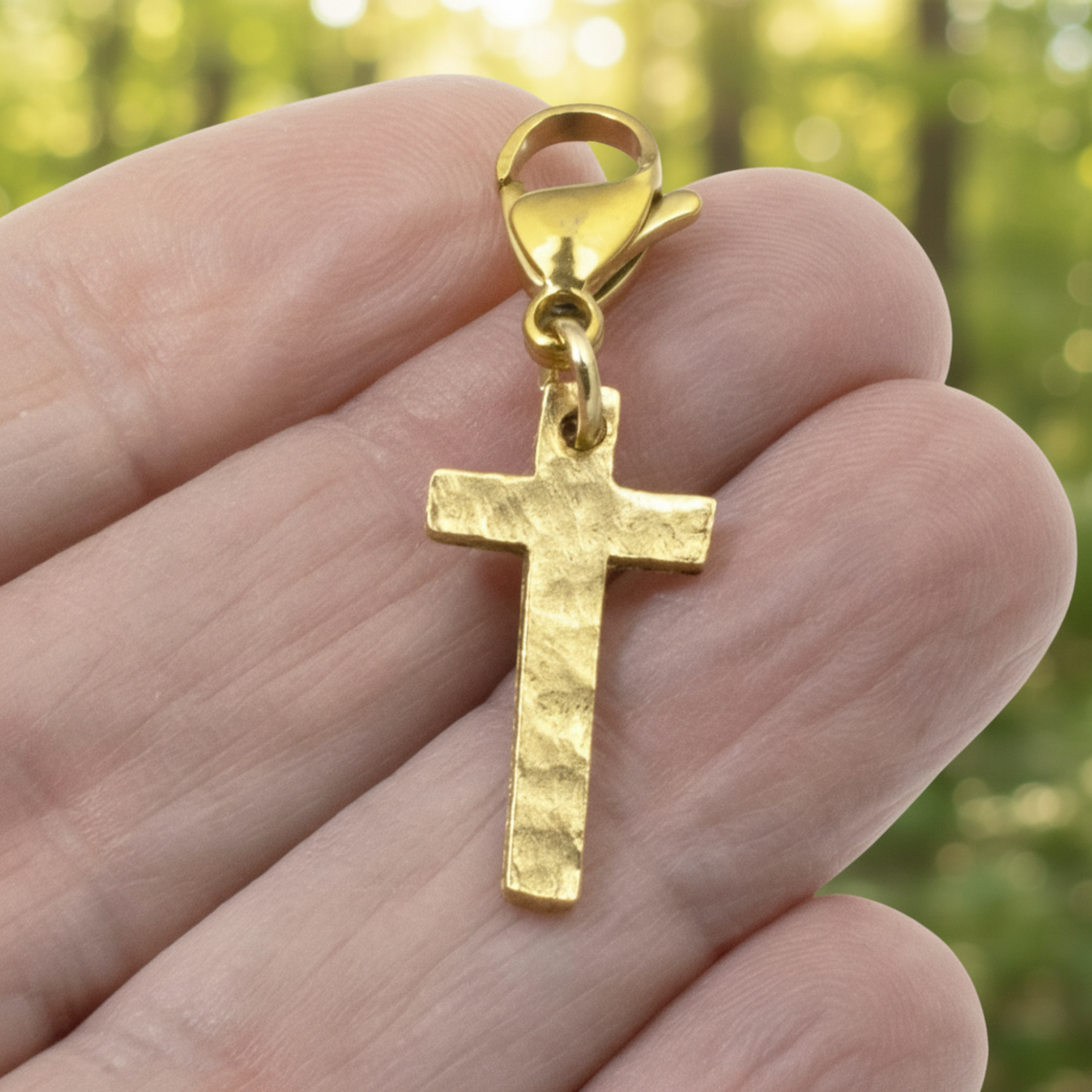 Hackberry Creek gold cross charm with a hammered texture and lobster clasp held in a hand against a blurred background