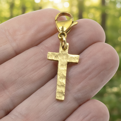 Hackberry Creek gold cross charm with a hammered texture and lobster clasp held in a hand against a blurred background