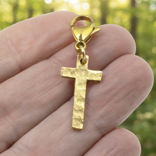 Hackberry Creek gold cross charm with a hammered texture and lobster clasp held in a hand against a blurred background