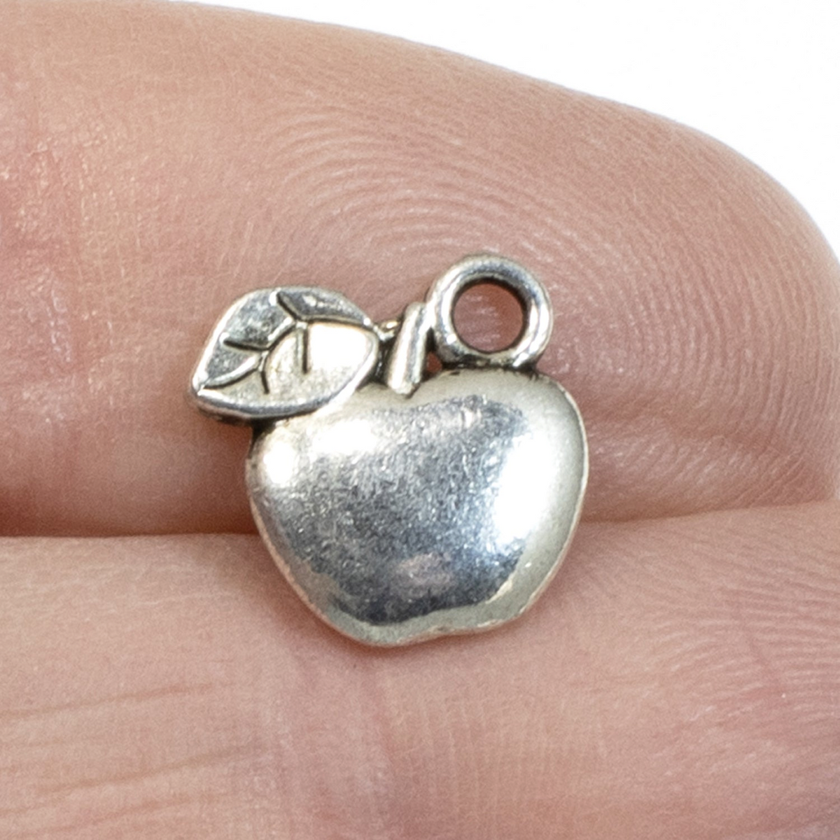 Hackberry Creek silver apple charm held on a finger, perfect for teacher jewelry and DIY craft projects.