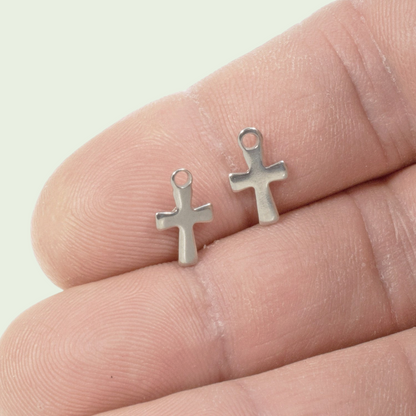 Hackberry Creek mini stainless steel cross charms held on a finger for scale, showing silver tone jewelry findings.