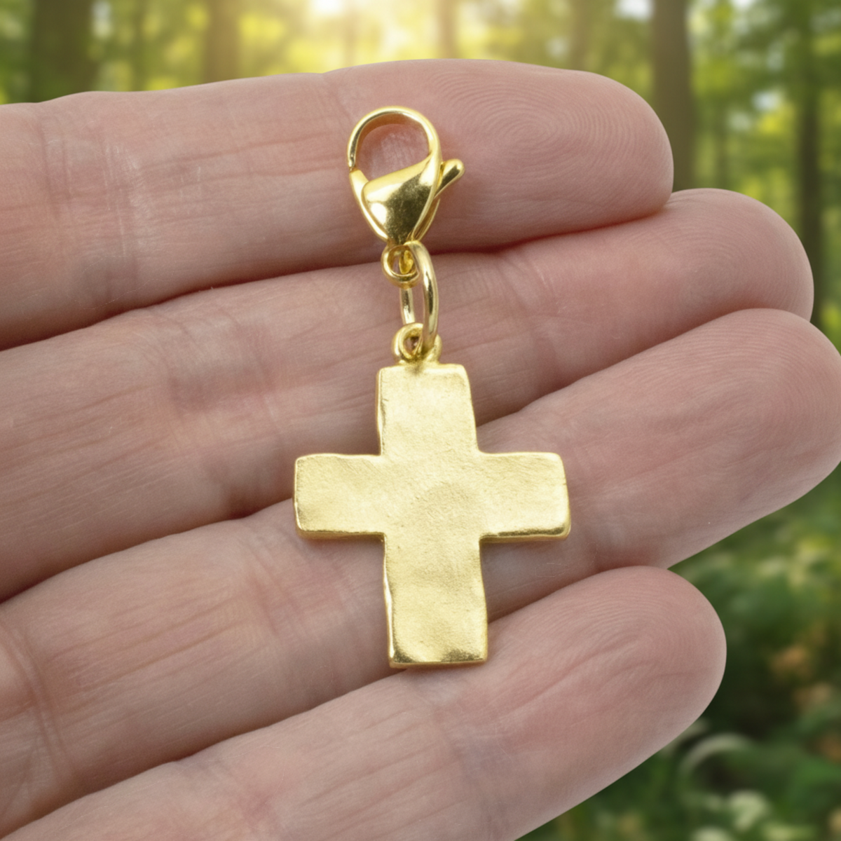 Hackberry Creek 24k gold-plated hammered cross charm with a lobster clasp held in a hand against a forest background.