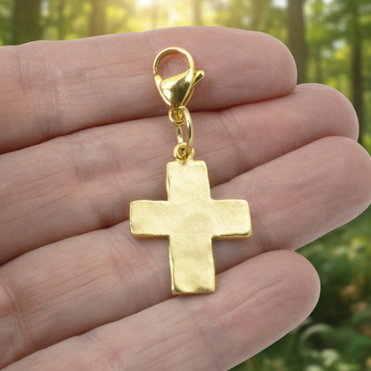 Hackberry Creek 24k gold-plated hammered cross charm with a lobster clasp held in a hand against a forest background.