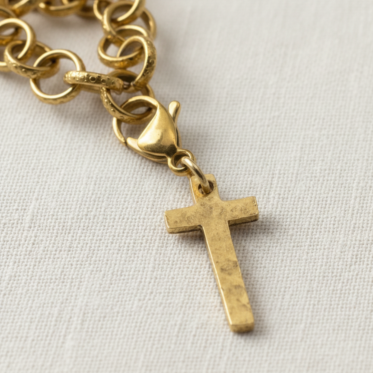Hackberry Creek gold cross charm with a hammered finish and lobster clasp attached to a gold chain on white fabric.