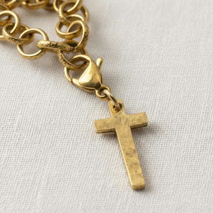 Hackberry Creek gold cross charm with a hammered finish and lobster clasp attached to a gold chain on white fabric.