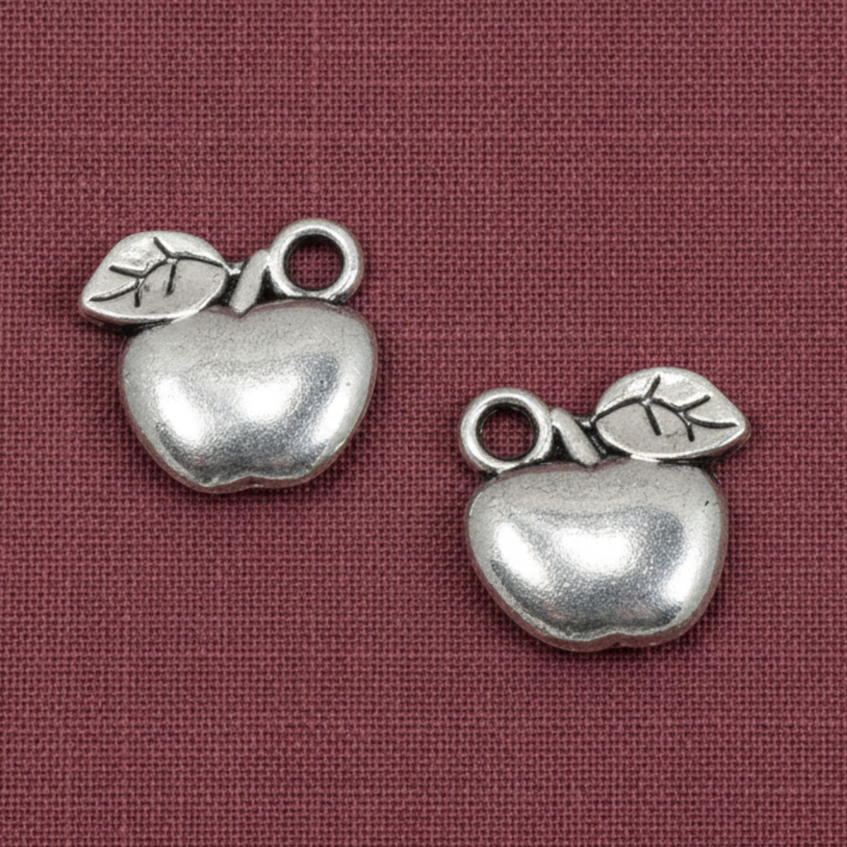 Hackberry Creek silver apple charms for jewelry making, featuring a durable zinc alloy design on a maroon background.