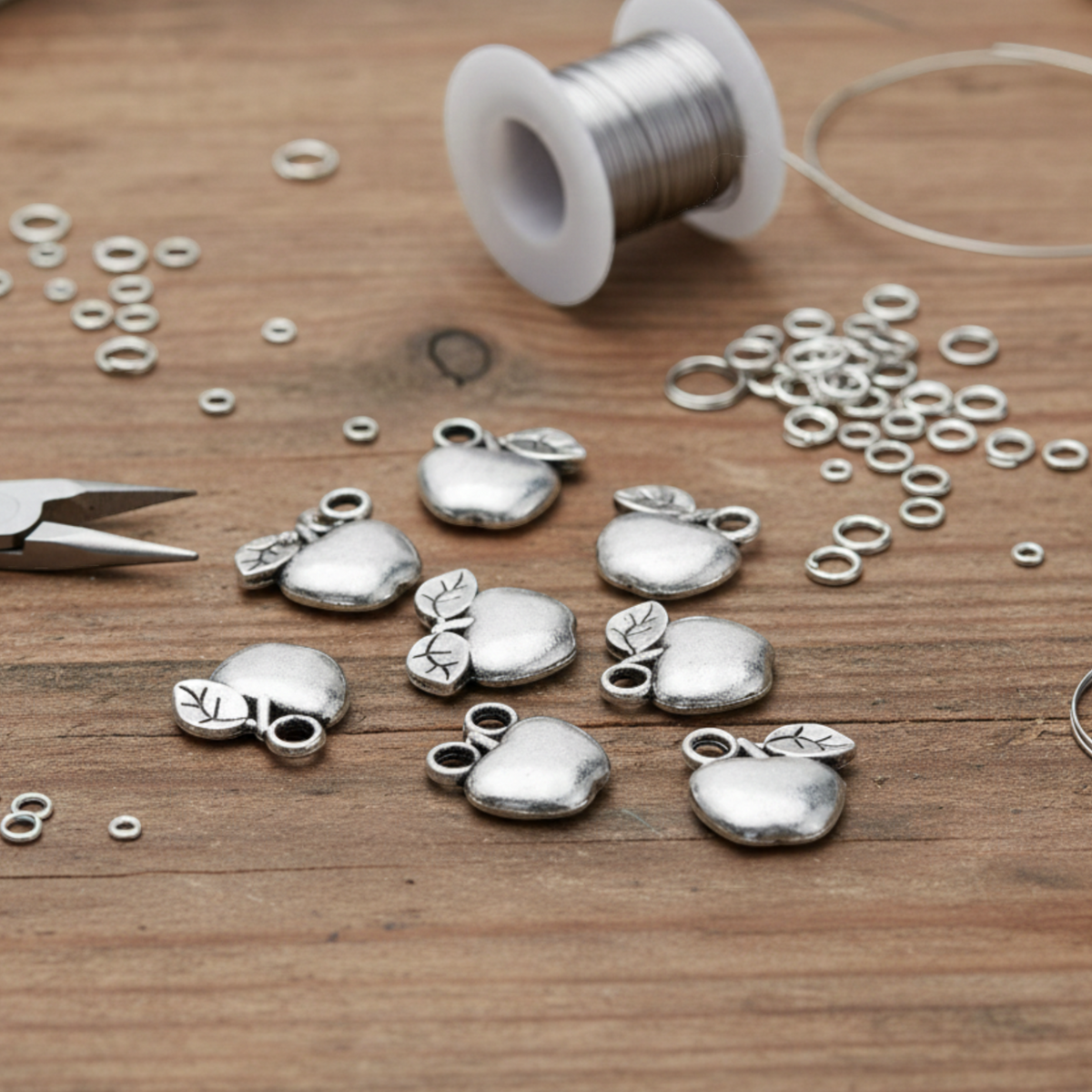 Hackberry Creek silver apple charms for jewelry making, displayed on a wooden table with crafting tools and wire.
