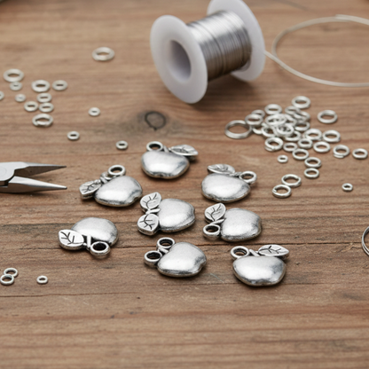 Hackberry Creek silver apple charms for jewelry making, displayed on a wooden table with crafting tools and wire.