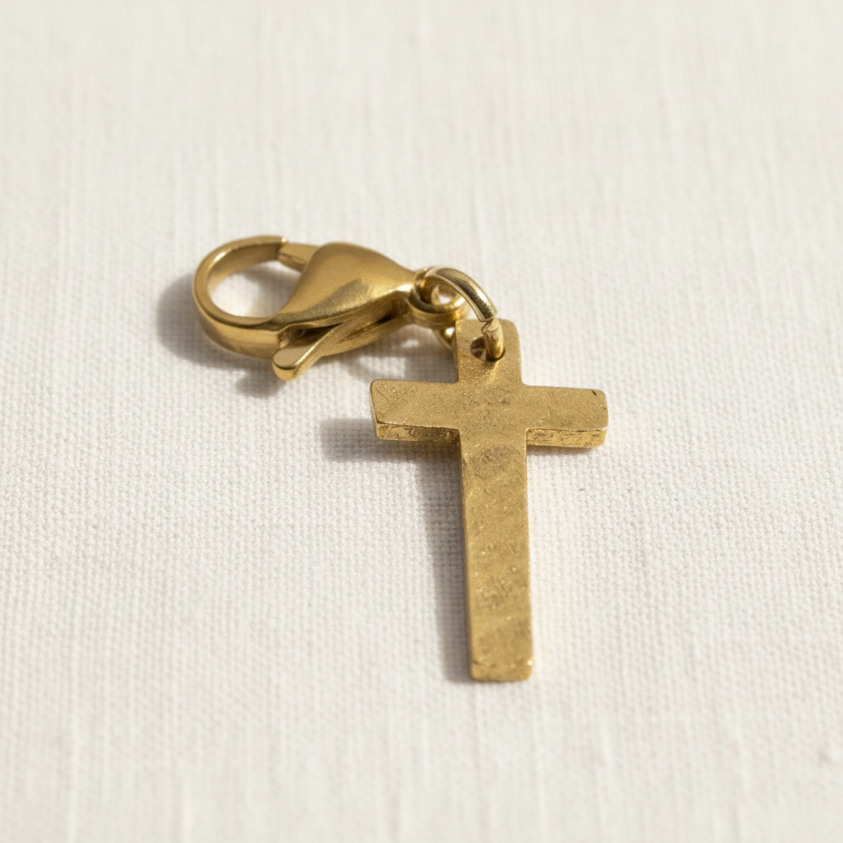 Hackberry Creek hammered gold cross charm with a stainless steel lobster clasp on a white fabric background