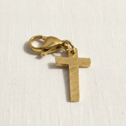 Hackberry Creek hammered gold cross charm with a stainless steel lobster clasp on a white fabric background