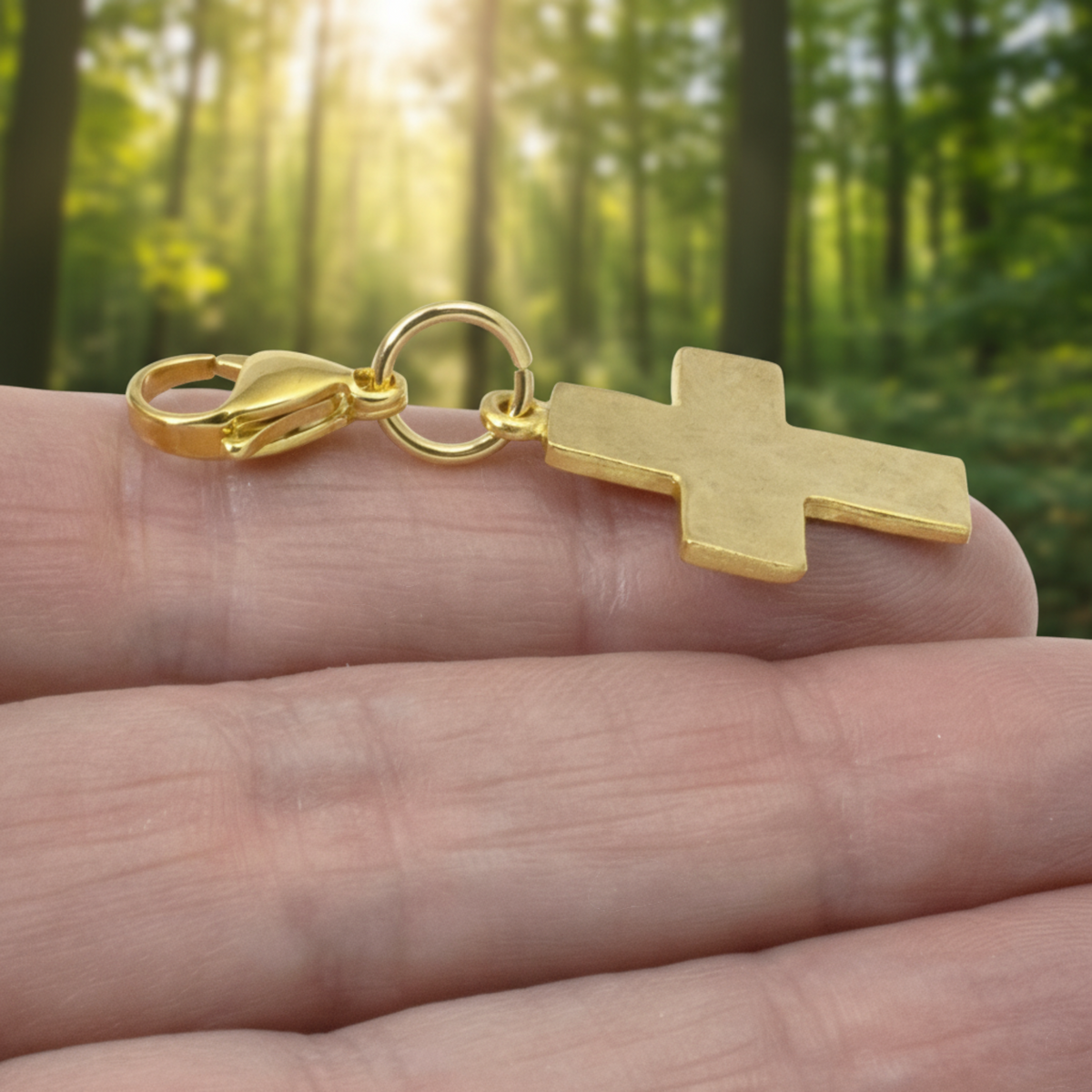Hackberry Creek gold cross clip-on charm with hammered texture held in a hand against a blurred forest background.