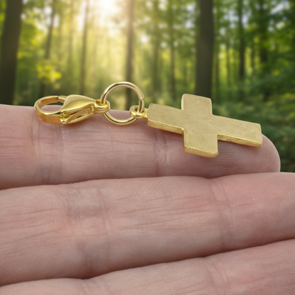 Hackberry Creek gold cross clip-on charm with hammered texture held in a hand against a blurred forest background.