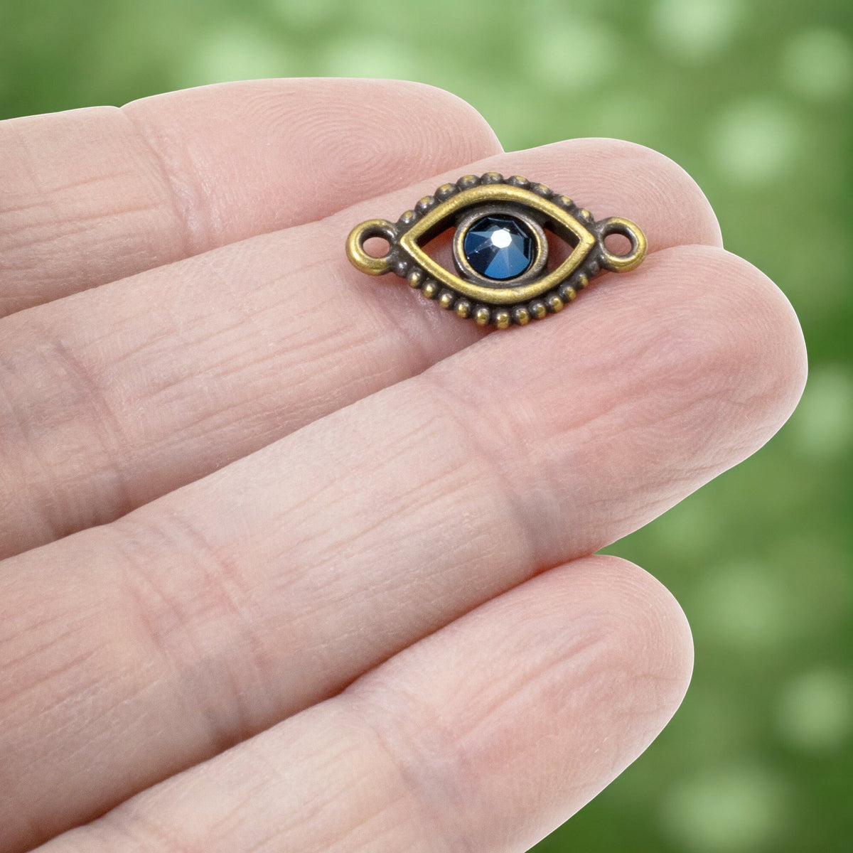 Hackberry Creek antique brass evil eye connector link with a blue crystal center held on a person's fingertip.