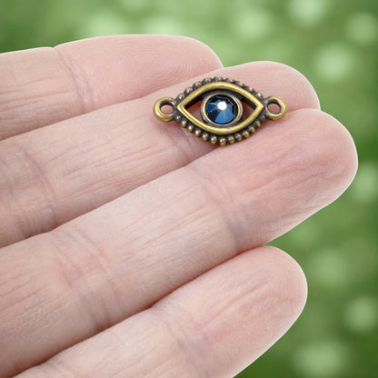 Hackberry Creek antique brass evil eye connector link with a blue crystal center held on a person's fingertip.