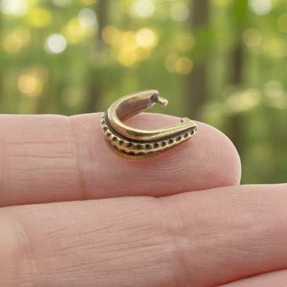 Hackberry Creek antique brass hammered pinch bail held on a finger with a blurred green forest background.