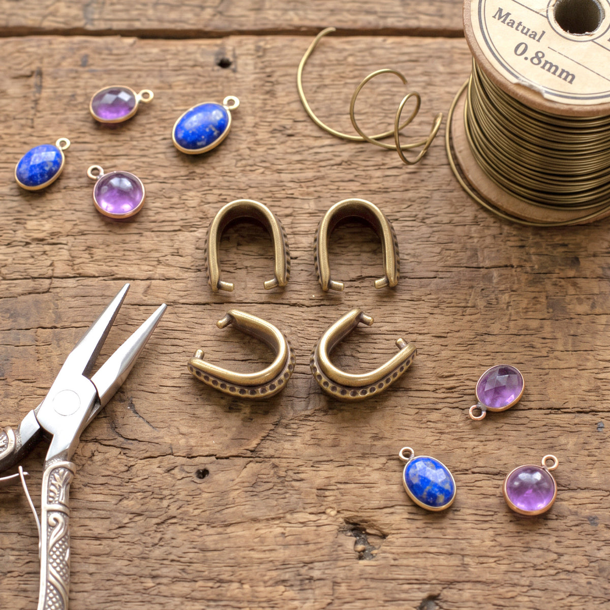 Hackberry Creek antique brass hammered pinch bails for jewelry making on a rustic wooden table with pliers and beads.