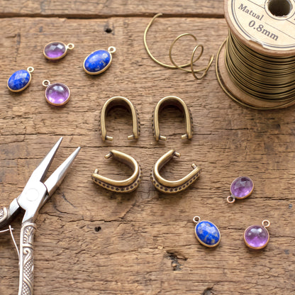Hackberry Creek antique brass hammered pinch bails for jewelry making on a rustic wooden table with pliers and beads.
