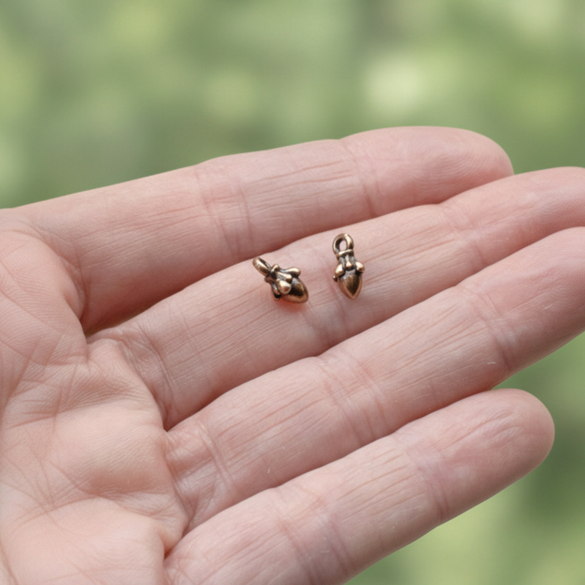 Hackberry Creek tiny antique copper acorn charms held in a palm for scale, showing 3D woodland jewelry detail.