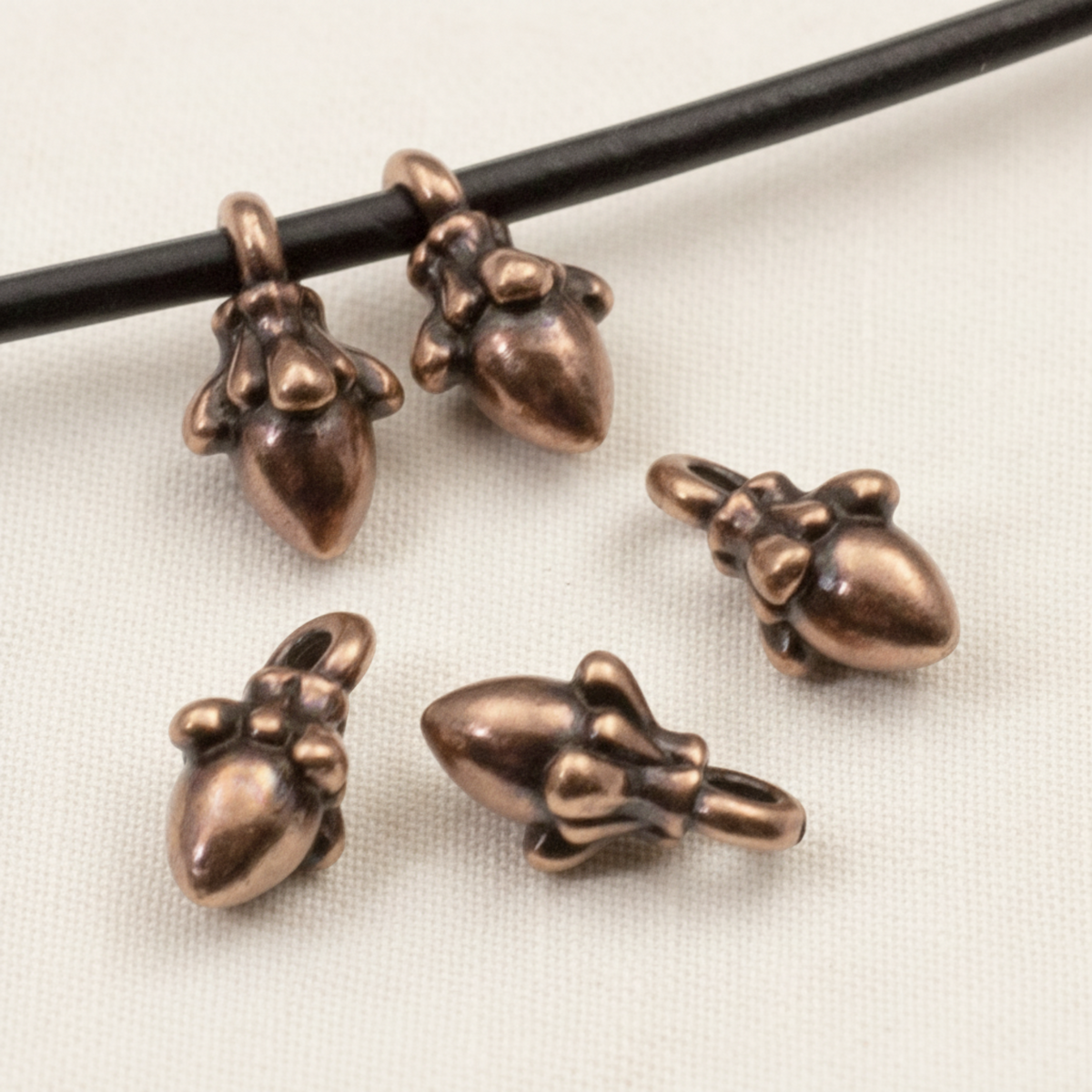 Hackberry Creek antique copper acorn charms with 3D sculpted detail, shown on a black leather cord for jewelry making.