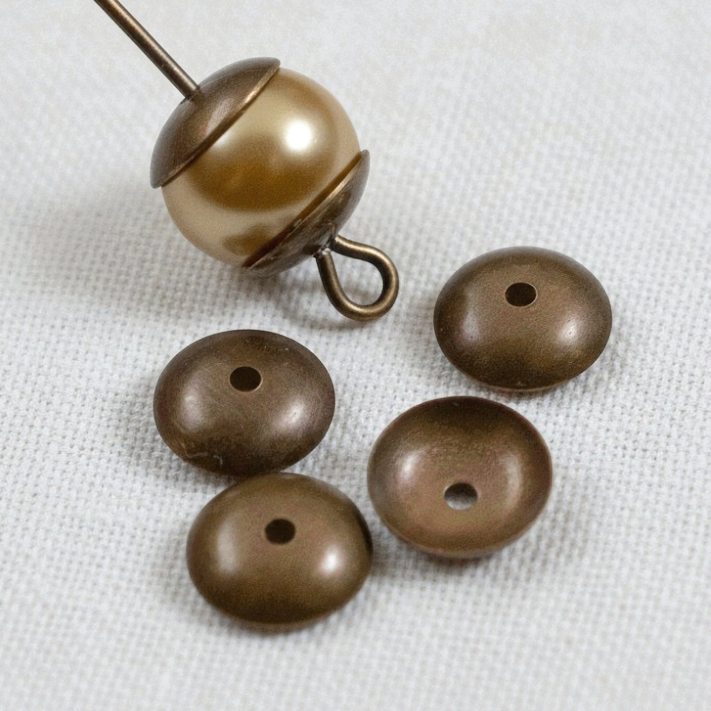 Hackberry Creek 7mm antiqued natural brass bead caps shown with a pearl bead on a headpin on a white fabric background.