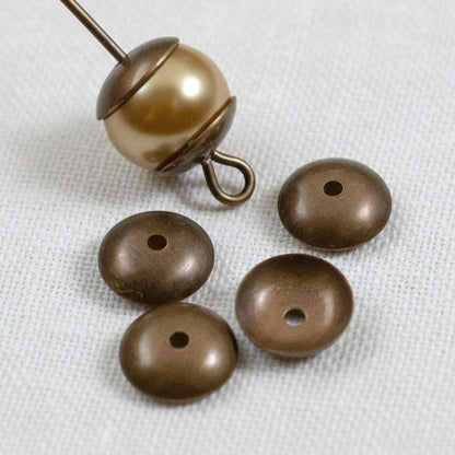 Hackberry Creek 7mm antiqued natural brass bead caps shown with a pearl bead on a headpin on a white fabric background.