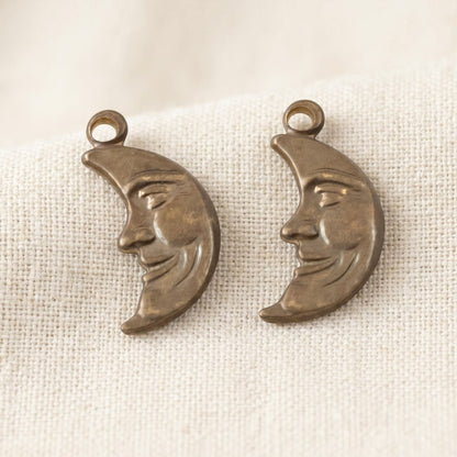 Hackberry Creek antiqued brass crescent moon charms with detailed man-in-the-moon faces on textured cream fabric.