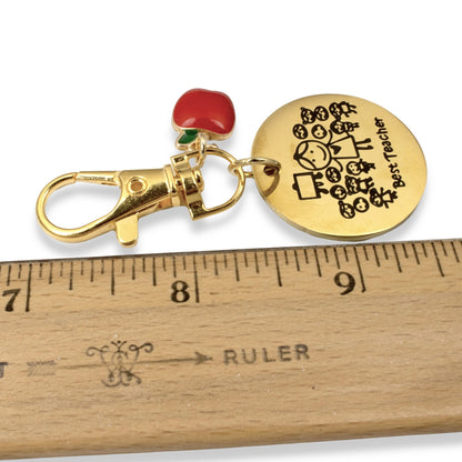 Hackberry Creek gold Best Teacher medallion charm with red enamel apple and swivel clasp next to a wooden ruler