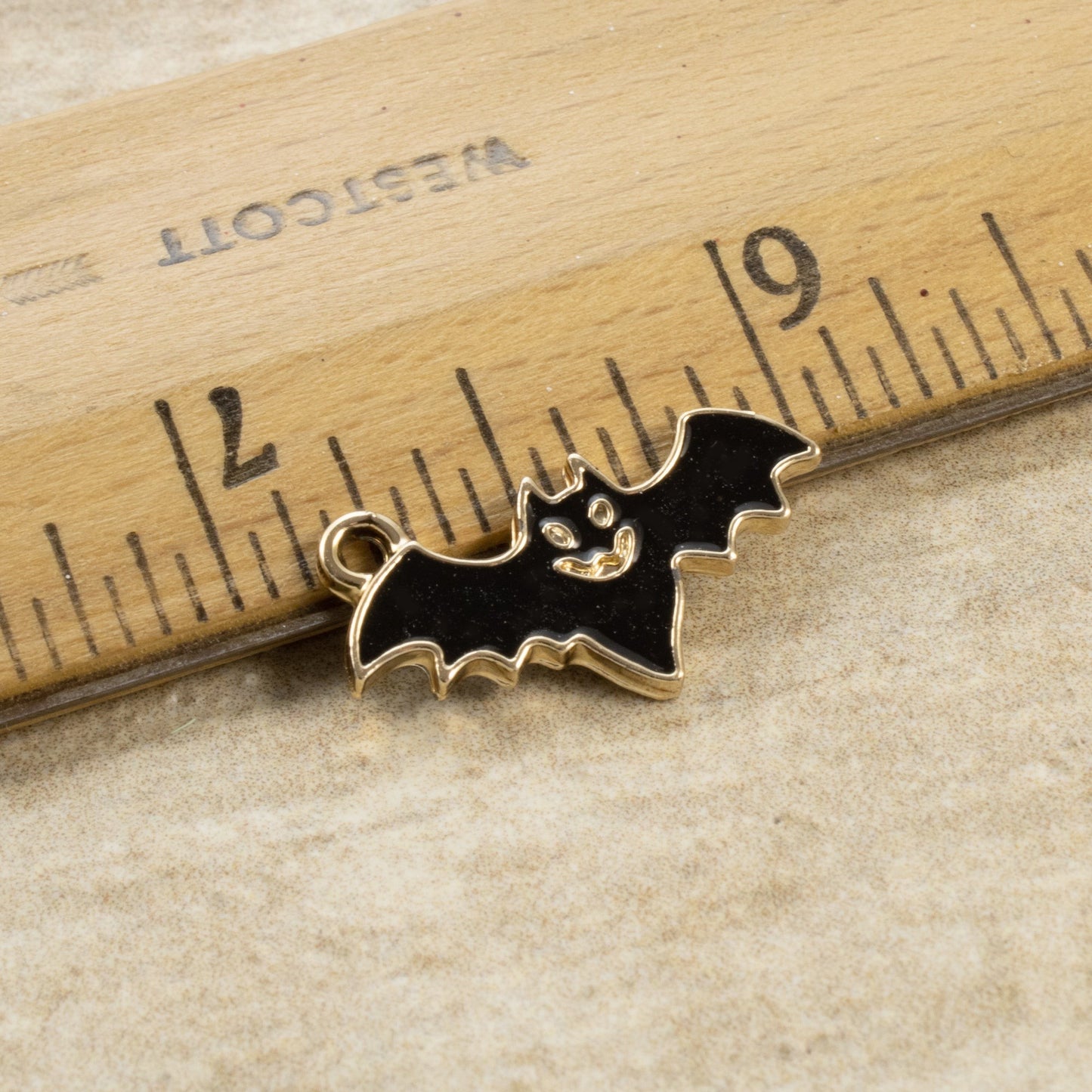 Hackberry Creek black enamel flying bat charm with gold trim next to a ruler for scale, ideal for gothic jewelry.