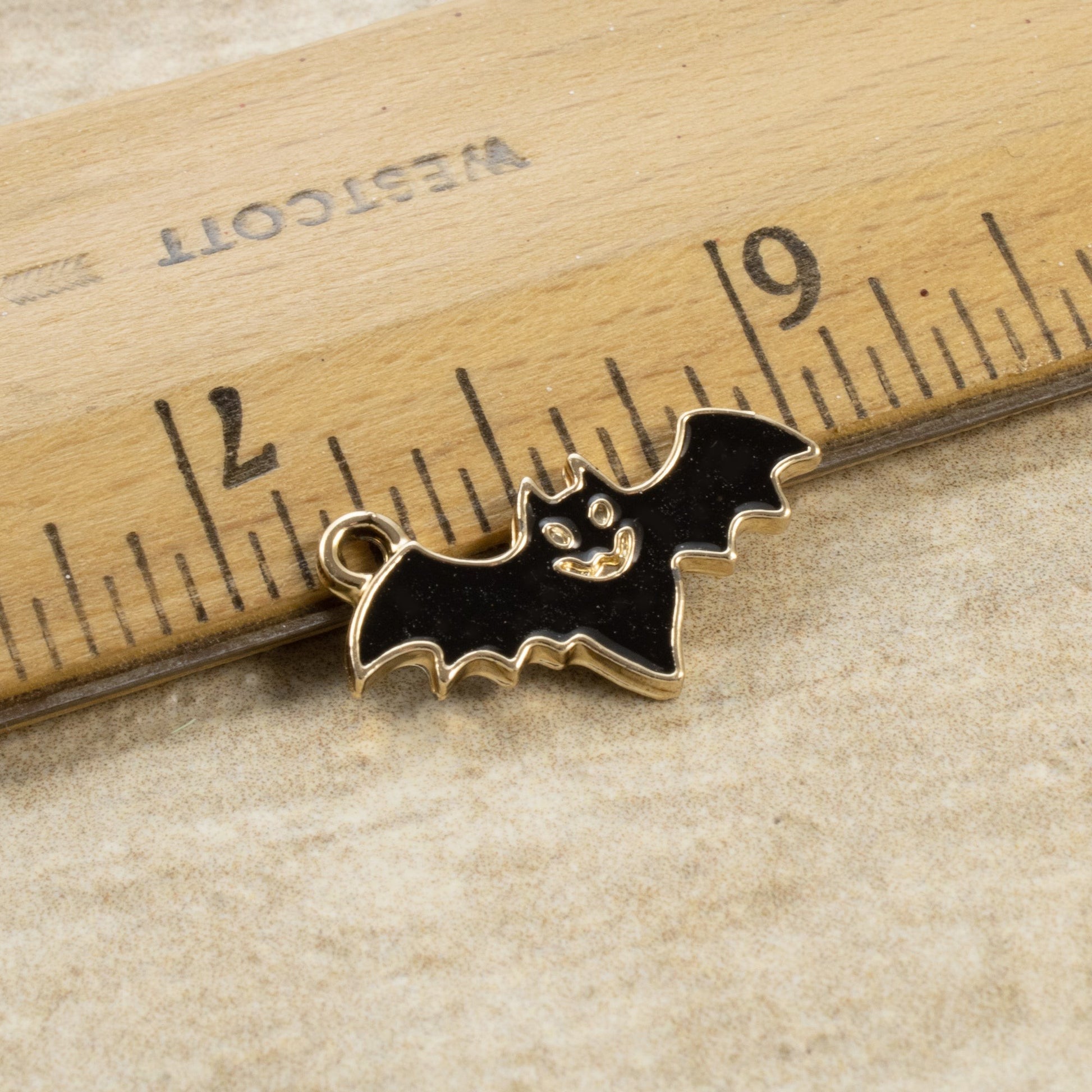 Hackberry Creek black enamel flying bat charm with gold trim next to a ruler for scale, ideal for gothic jewelry.