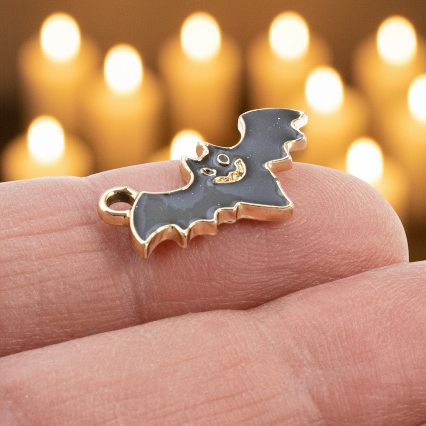 Hackberry Creek black enamel flying bat charm held on a finger with a blurred candle background for gothic jewelry