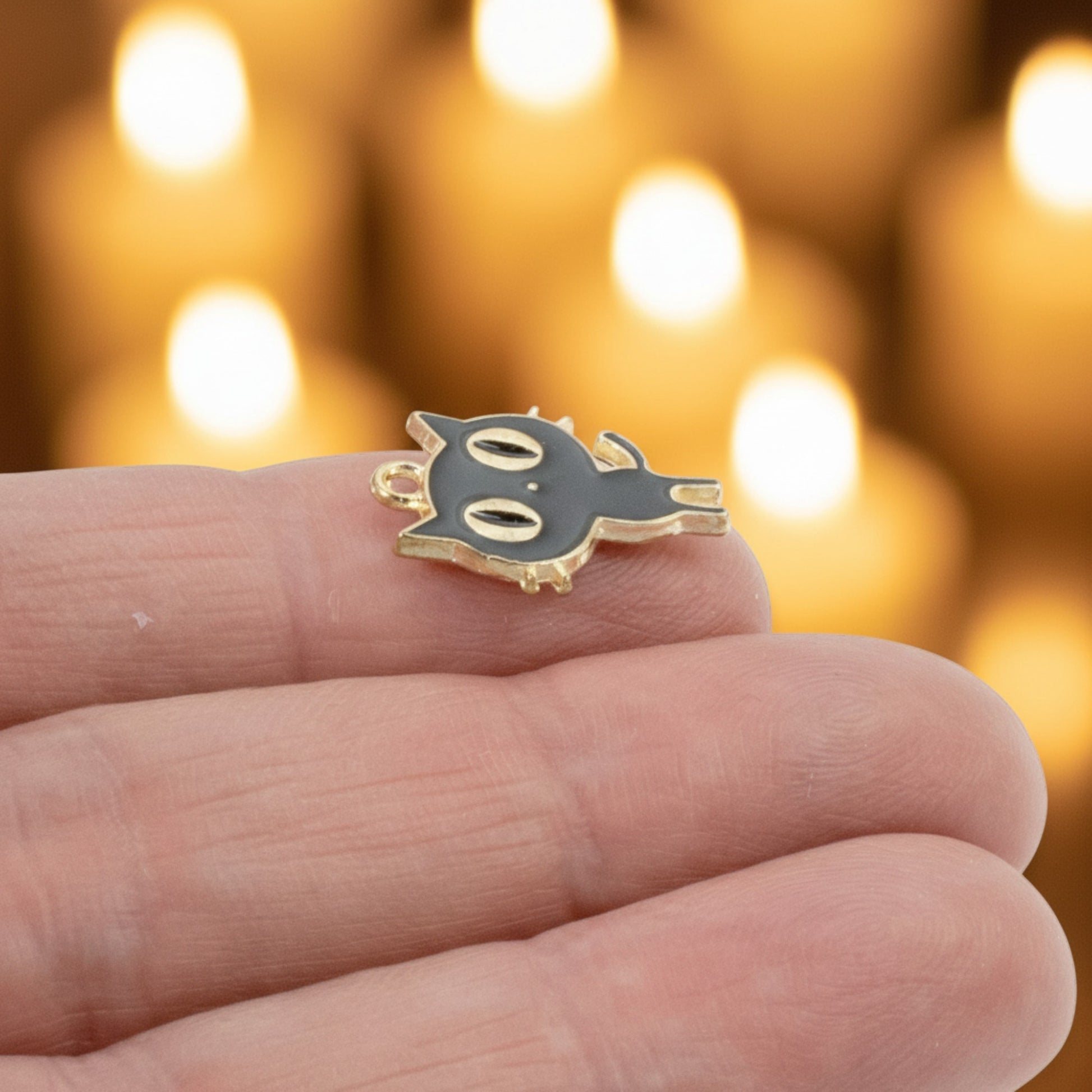Hackberry Creek black enamel cat charm with gold plating held between fingers against a blurred candle background.