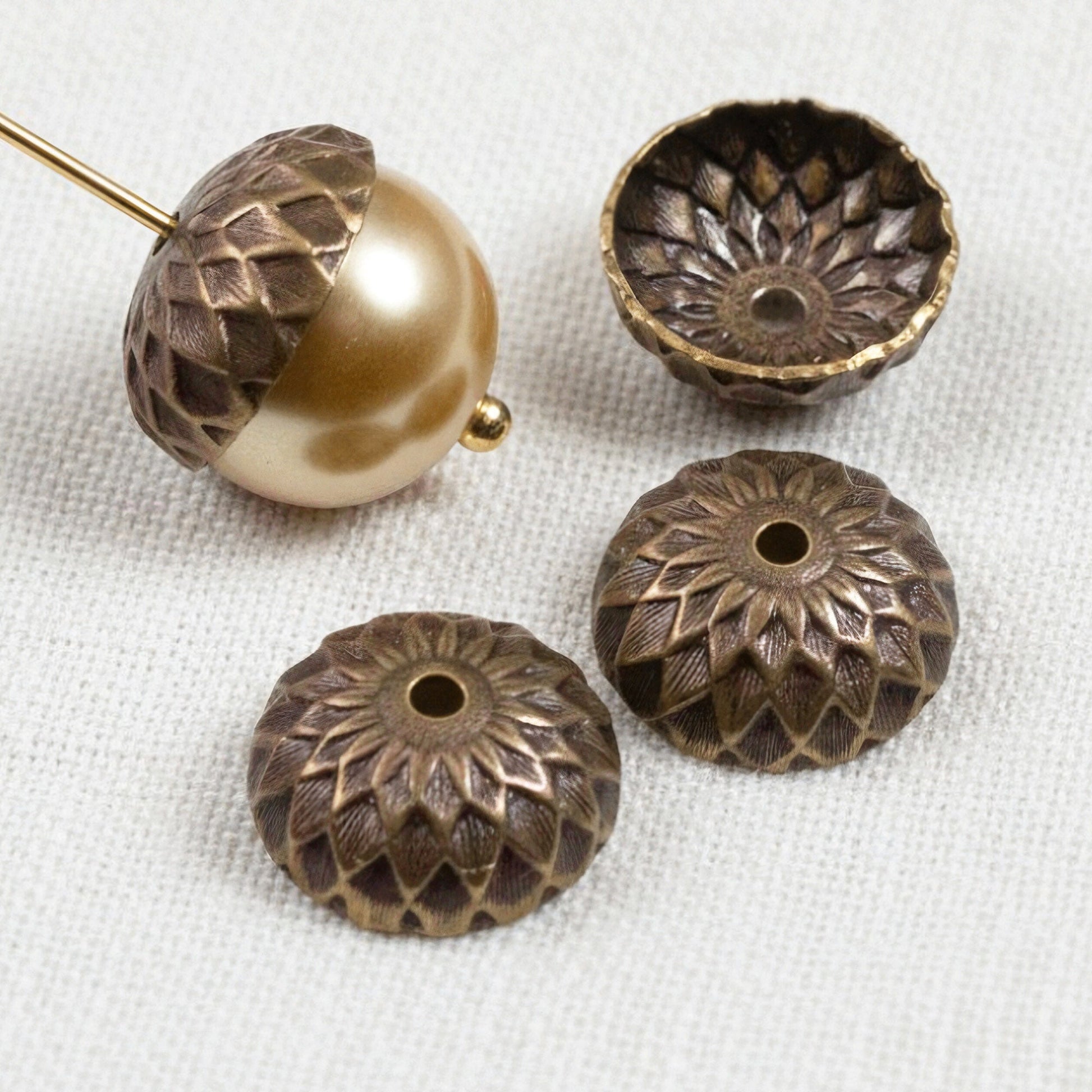Hackberry Creek 13mm natural brass acorn bead caps with detailed sunflower relief stamping on a white fabric background.