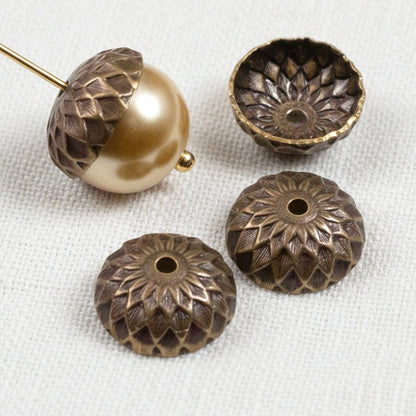 Hackberry Creek 13mm natural brass acorn bead caps with detailed sunflower relief stamping on a white fabric background.