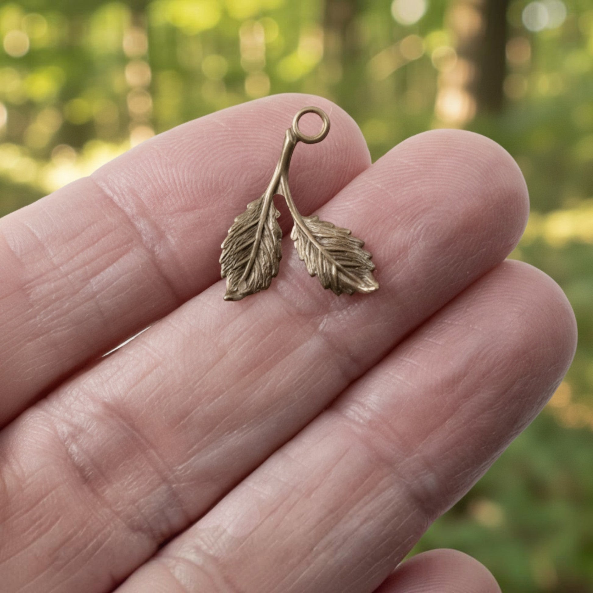 Hackberry Creek antiqued brass double-sided beech leaf charm held on a finger against a blurred forest background.