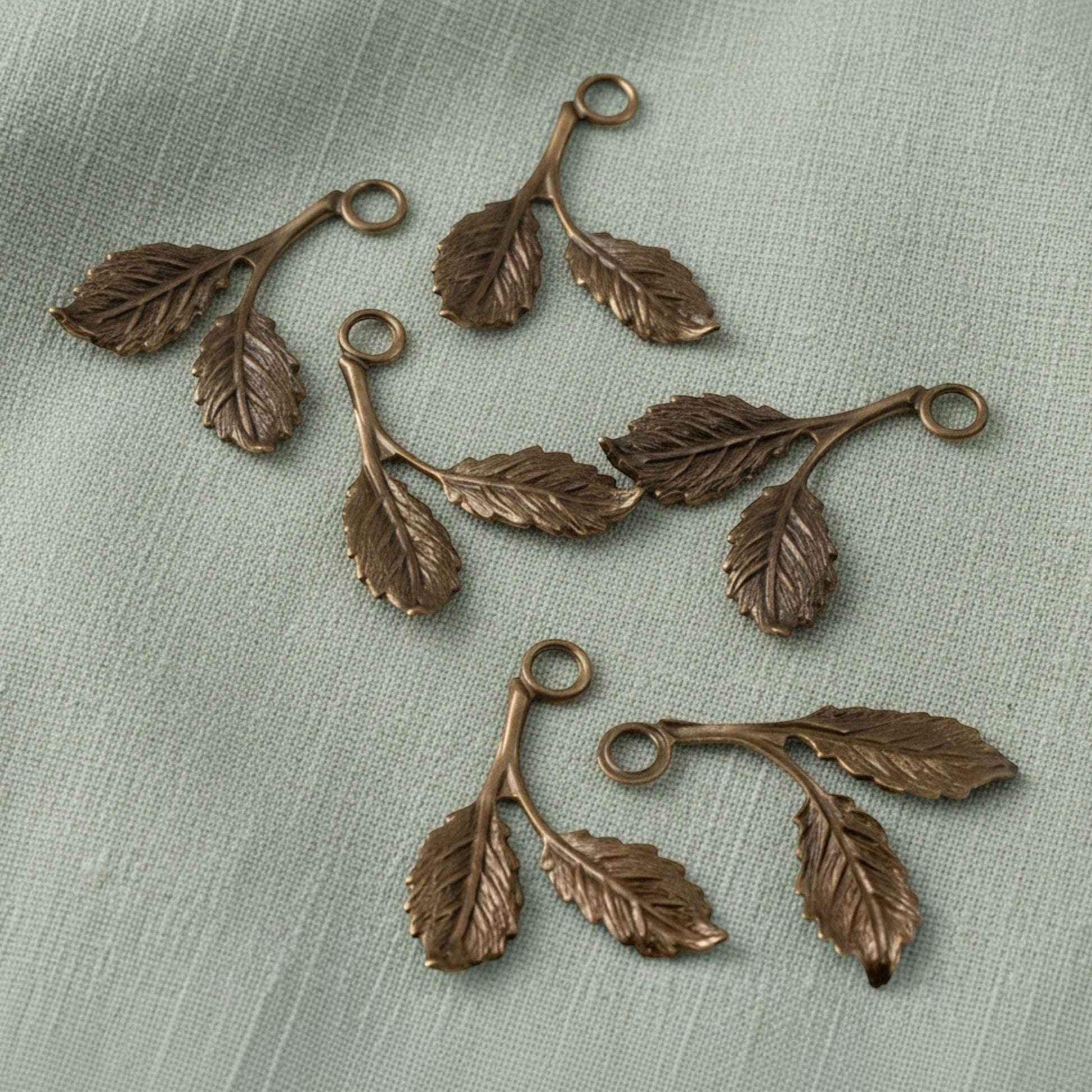 Hackberry Creek antiqued brass beech leaf charms with detailed botanical texture on a sage green fabric background.