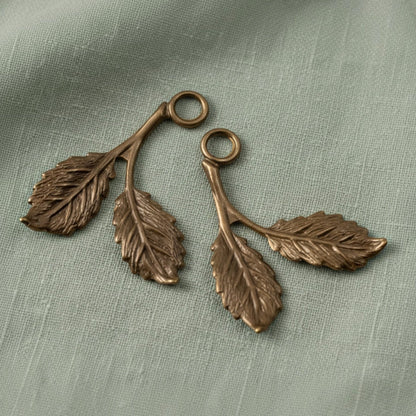 Hackberry Creek antiqued brass beech leaf charms with detailed botanical texture on a sage green fabric background.