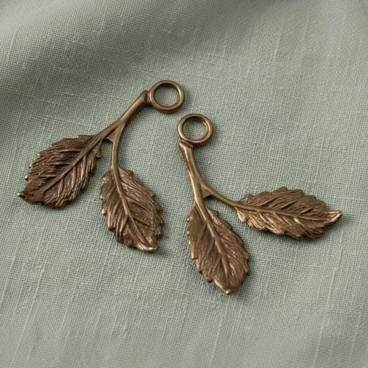Hackberry Creek antiqued brass beech leaf charms with detailed botanical texture on a sage green fabric background.