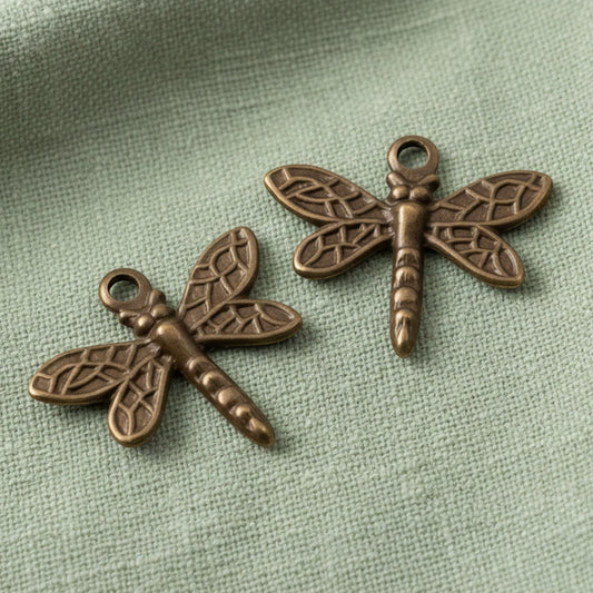 Hackberry Creek antiqued brass dragonfly charms with detailed wing patterns on a sage green fabric background.