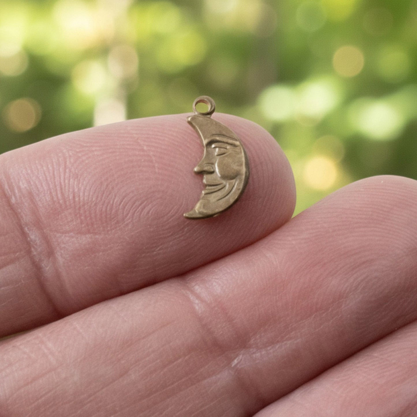 Hackberry Creek antiqued brass crescent moon charm with a detailed face held on a fingertip for scale.