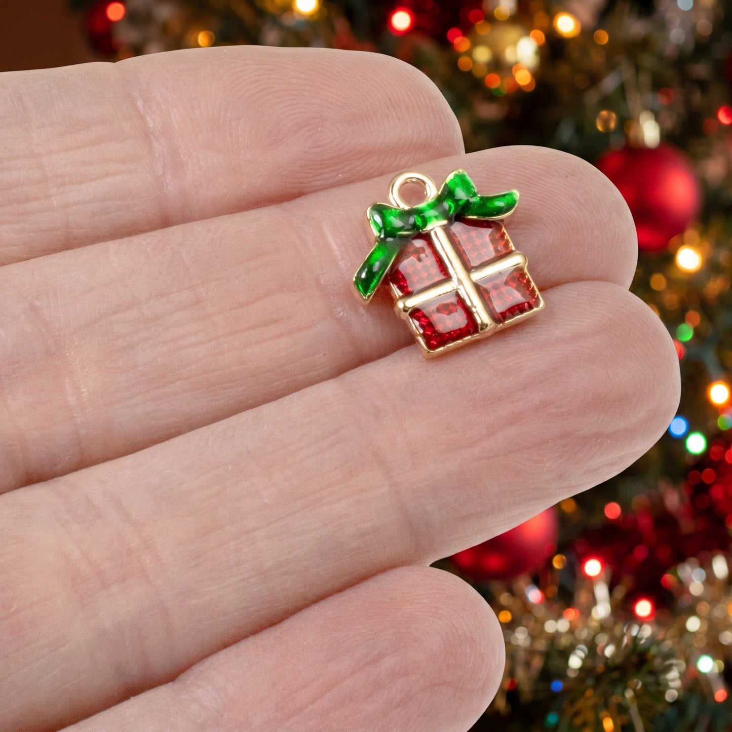 Hackberry Creek Christmas present charm with red enamel and green bow, held in hand against a festive tree background.