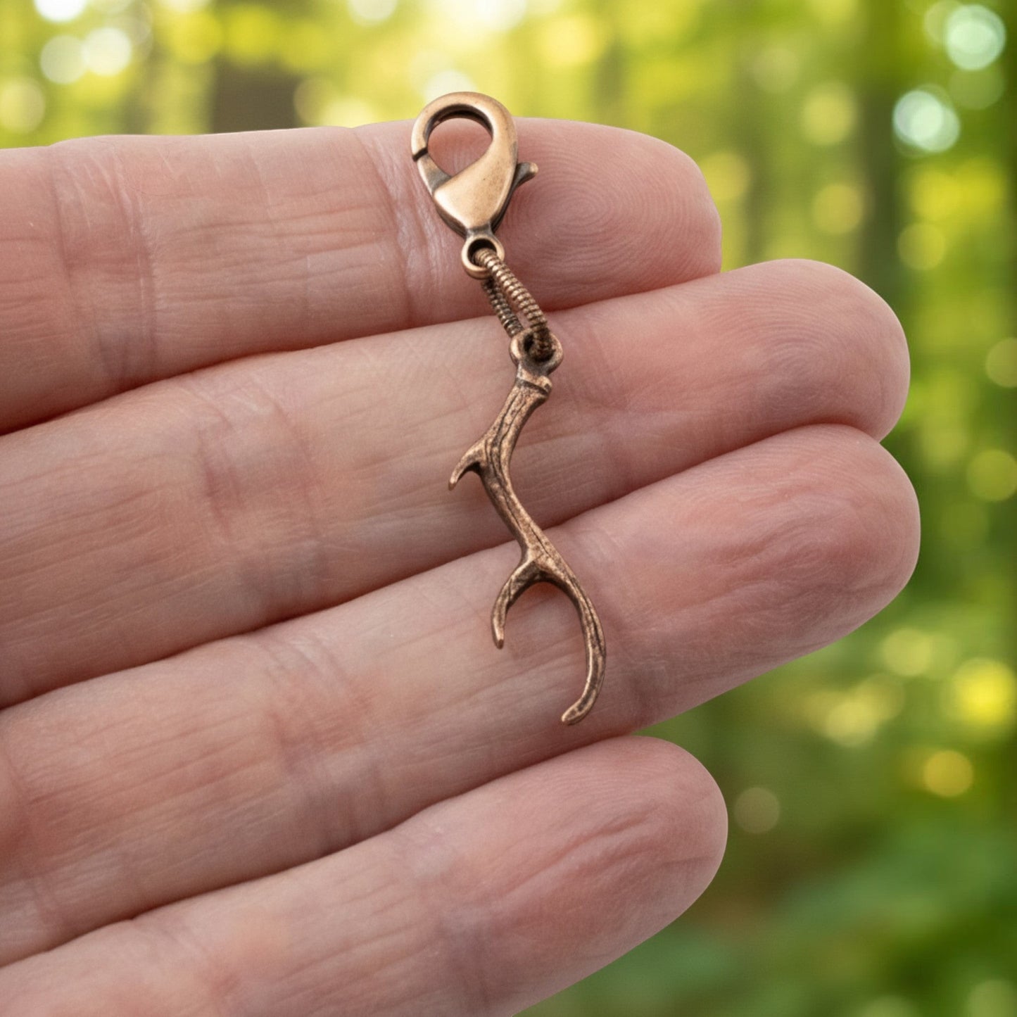 Hackberry Creek copper antler clip-on charm held in a hand against a blurred woodland background.