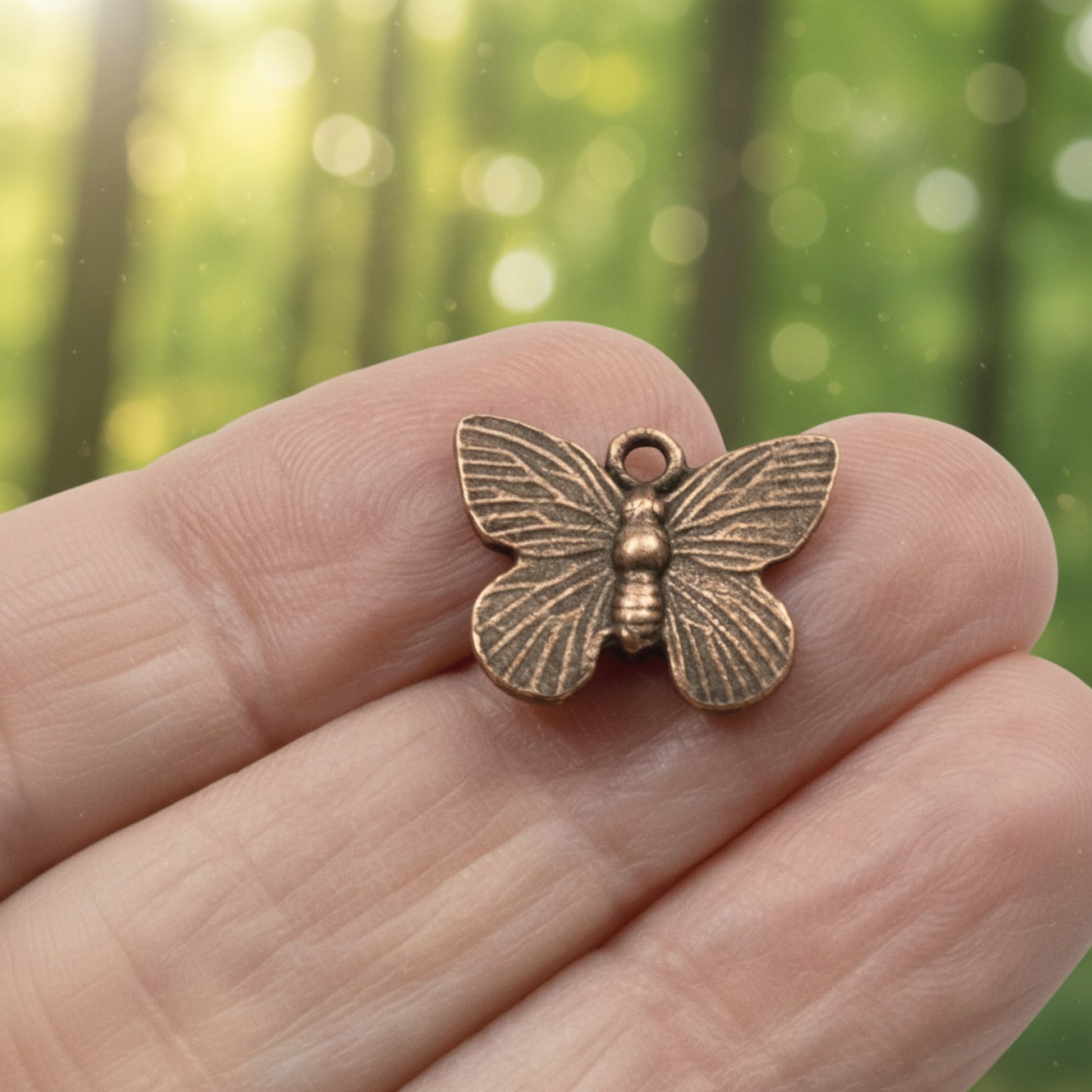 Hackberry Creek antique copper butterfly charm held in hand against a soft-focus forest background.