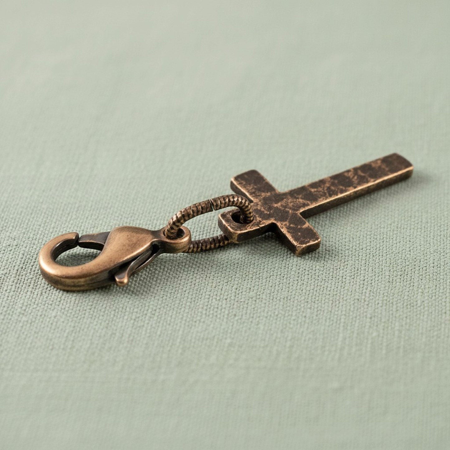 Hackberry Creek narrow copper cross charm with distressed patina and lobster clasp on a light green fabric background.