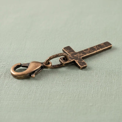 Hackberry Creek narrow copper cross charm with distressed patina and lobster clasp on a light green fabric background.