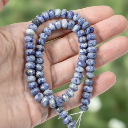 Hackberry Creek faceted denim lapis rondelle beads in blue and gray held in a hand against a blurred green background.